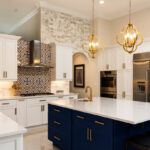 Modern kitchen with white cabinets, blue island, gold hardware, stainless steel appliances, patterned tile backsplash, and two pendant lights over the island.