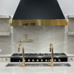 Modern kitchen with a marble island, double sinks, gold fixtures, a large black and gold range hood, marble backsplash, and built-in shelving; staircase visible on the right.